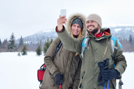 Cheerful active couple in warm clothing hiking outdoors with backpacks in winter mountains and taking selfie on phoneの写真素材