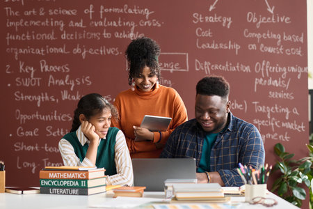 Front view portrait of three Black college students looking at laptop screen against blackboard with writings in backgroundの写真素材