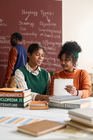 Vertical portrait of two young Black girls using digital tablet together while studying in school classroomの写真素材