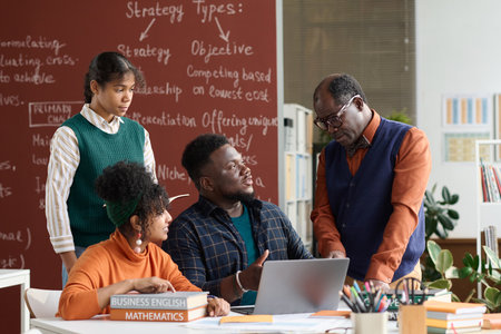 Portrait of senior African American teacher talking to group of students sitting at table with books and computerの写真素材