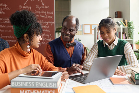 Portrait of smiling senior professor talking to two African American students sitting at table with laptop in college classroomの写真素材