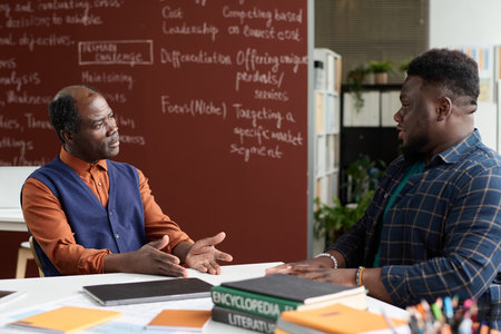 Portrait of Black senior professor listening to male student during one on one consultation in school classroomの写真素材