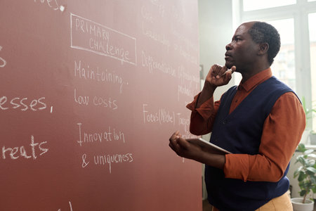 Side view portrait of senior Black college professor thinking by chalkboard while preparing for lecture in collegeの写真素材