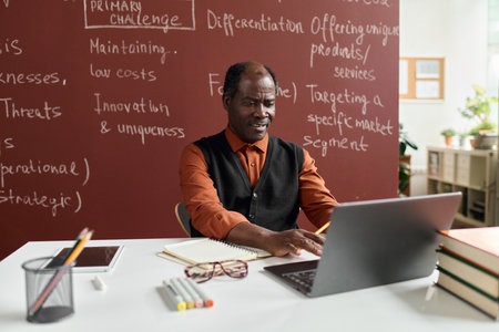 Portrait of senior African American college professor sitting at workplace in college and using laptopの写真素材