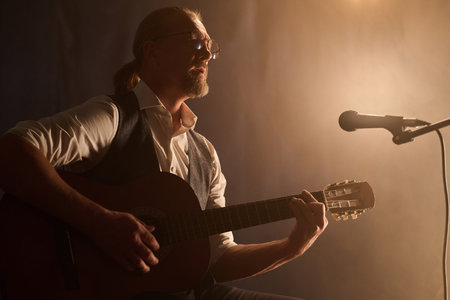 Side view portrait of classic mature guitarist playing acoustic guitar during stage performance in soft spotlight, copy spaceの写真素材