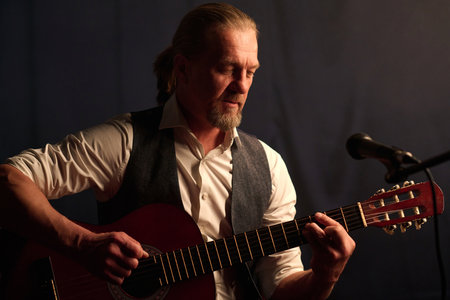 Portrait of classic mature musician playing acoustic guitar during stage performance and singing to microphoneの写真素材