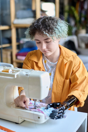 Vertical portrait of teenage girl with prosthetic arm using sewing machine in inclusive atelier studioの写真素材