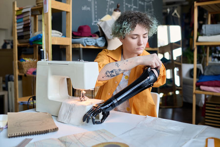 Portrait of teenage girl adjusting prosthetic arm while working in inclusive atelier studio and sewing clothing copy spaceの写真素材