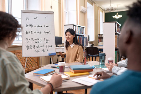 Portrait of young Asian woman as female teacher talking to group of students sitting at table by whiteboard with hieroglyphs copy spaceの写真素材