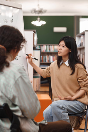 Vertical portrait of young Asian woman explaining grammar rules of Chinese language to students in library and pointing at boardの写真素材
