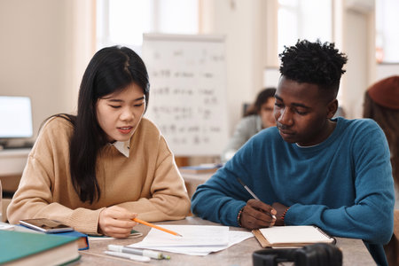 Front view portrait of two multiethnic students Asian woman and Black man studying together sitting at table in college libraryの写真素材