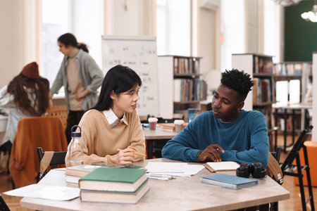 Portrait of young Asian woman and Black man sitting at table in class together during pair studyの写真素材
