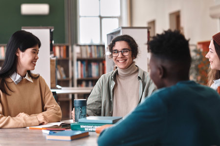 Portrait of young male student with genuine smile enjoying schoolwork sitting at table with friends in libraryの写真素材