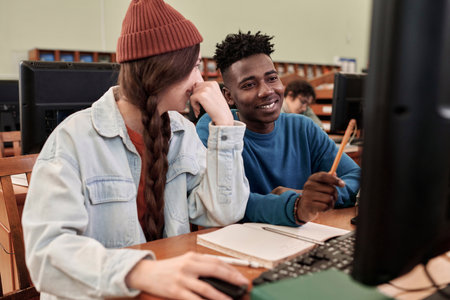 Portrait of two young students using computer in college library and pointing at screen while working on project togetherの写真素材