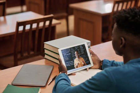 Over shoulder view of young Black man watching video lecture in school library and using digital tablet with teacher speaking on screenの写真素材
