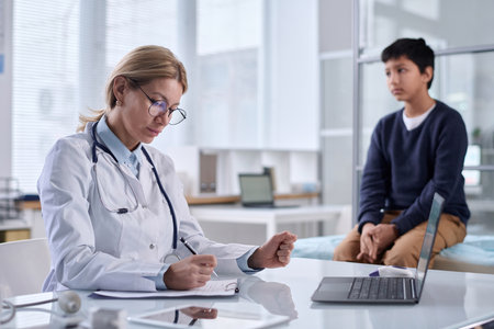 Portrait of woman doctor wearing glasses taking notes with child sitting on cot in background copy spaceの写真素材