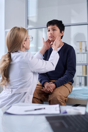 Vertical portrait of pediatrician examining neck and throat of young boy during health check up in clinicの写真素材