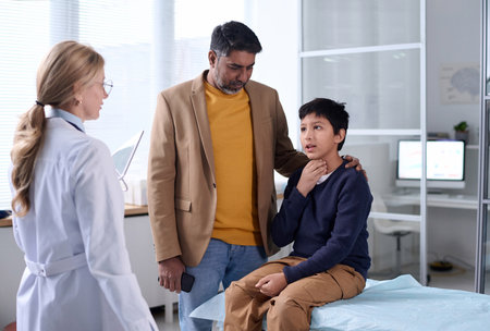 Portrait of Middle Eastern father and son visiting doctor in clinic with young boy explaining symptoms and pointing at throatの写真素材