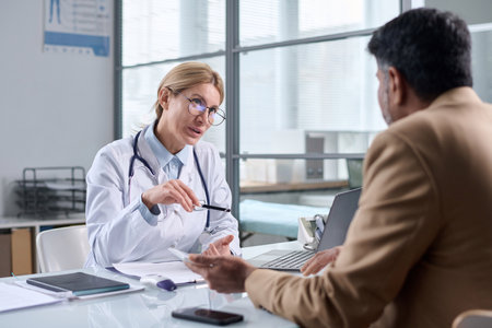 Portrait of mature female doctor wearing glasses and talking to patient across table in medical clinicの写真素材