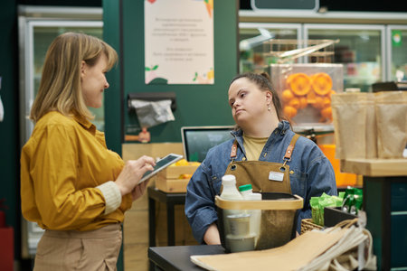 Waist up portrait of young woman with disability working in supermarket with manager assisting and giving instructionsの写真素材