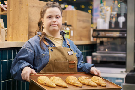 Waist up portrait of confident young woman with Down syndrome working in bakery and looking at camera holding tray with fresh pastry copy spaceの写真素材