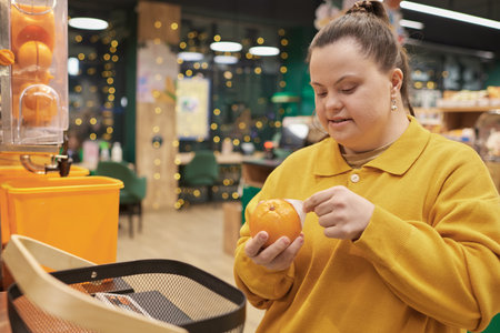 Portrait of cheerful young woman with Down syndrome buying fresh oranges in fruit section of supermarket and wearing yellowの写真素材