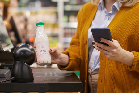 Close up of unrecognizable woman scanning milk bottle while using self checkout service grocery shopping and using smartphonの写真素材