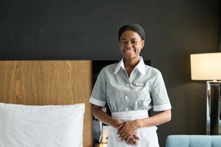 Waist up portrait of young Black woman as housekeeper wearing uniform and smiling at camera standing in hotel room copy spaceの写真素材