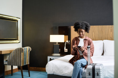 Wide angle view at young Black woman sitting on bed in hotel room and using smartphone with suitcase copy spaceの写真素材