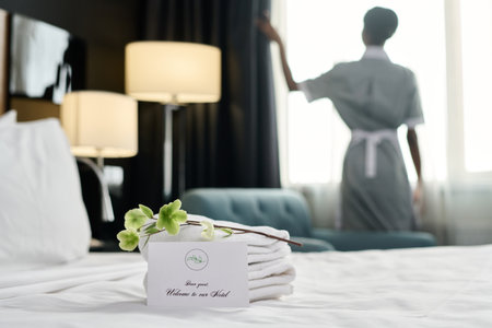Welcome note with delicate flower decor on bed in luxury in hotel room and blurred silhouette of housekeeper copy spaceの写真素材