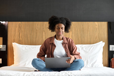 Portrait of young African American woman using laptop in hotel room while sitting on bed with legs crossed copy spaceの写真素材