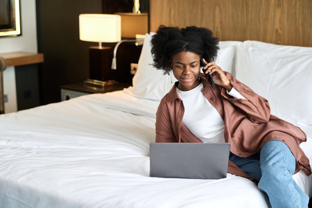 Portrait of young African American woman using laptop and phone on bed in hotel room during business travel copy spaceの写真素材