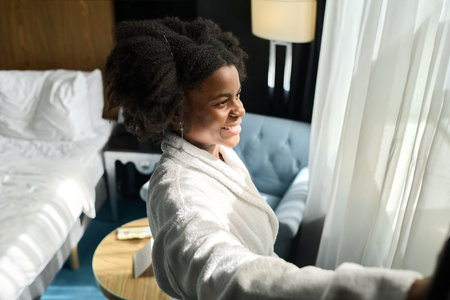 Side view portrait of happy young woman wearing bathrobe enjoying morning in hotel room and opening curtainsの写真素材