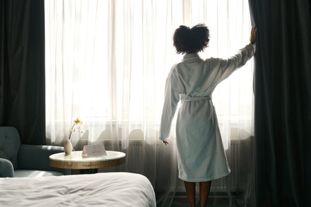 Back view of African American woman wearing bathrobe enjoying morning in hotel room and opening window curtains, copy spaceの写真素材