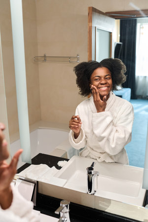 Vertical portrait of young African American woman looking in mirror enjoying skincare in morning, copy spaceの写真素材