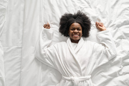 Minimal top view of young Black woman falling onto bed enjoying relaxing SPA vacation in hotel and smiling at camera, copy spaceの写真素材
