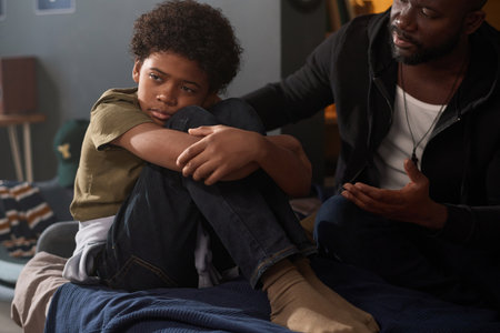 Portrait of depressed young boy sitting on bed and hugging knees with caring father talking to son affectionately copy spaceの写真素材
