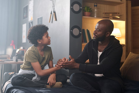 Side view portrait of caring African American father talking to son and holding hands sitting on bed togetherの写真素材