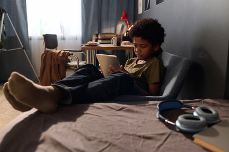 Full length portrait of African American teenage boy using digital tablet sitting on bed in dark room copy spaceの写真素材