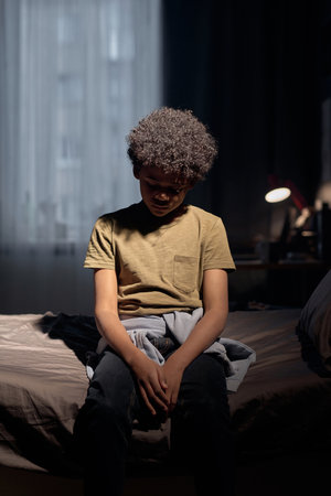 Vertical portrait of young Black boy sitting on bed with dark dramatic lighting lonely and upsetの写真素材