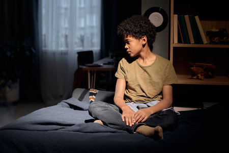 Cinematic side view portrait of teenage African American boy sitting on bed with dramatic light and shadow copy spaceの写真素材