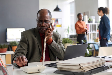 Front view portrait of senior man using landline phone at workplace in age inclusive office space with young people in background copy spaceの写真素材