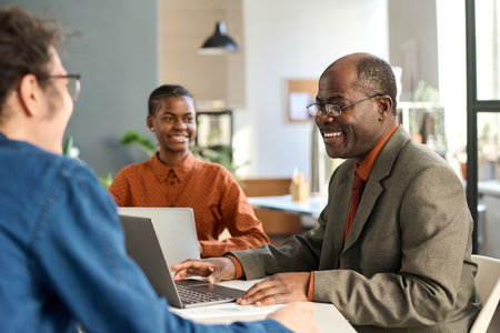 Side view portrait of Black senior man laughing enjoying work with young colleagues in office during business meetingの写真素材