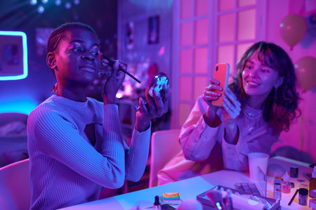 Portrait of two young women using luminescent makeup in neon room and taking photos with phoneの写真素材