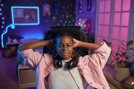 Front view portrait of young Black woman relaxing in gaming chair at night and wearing headphonesの写真素材