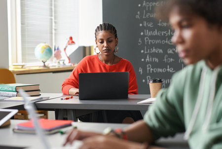 Portrait of Black young woman using laptop in school classroom with defocused students in...