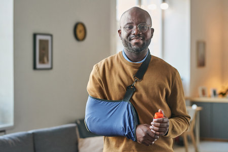 Waist up portrait of smiling adult African American man with broken arm in sling looking at camera and holding bottle of pills copy spaceの写真素材