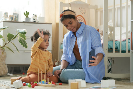 Woman spending quality time with young child, sitting on floor in nursery room filled with toys and...