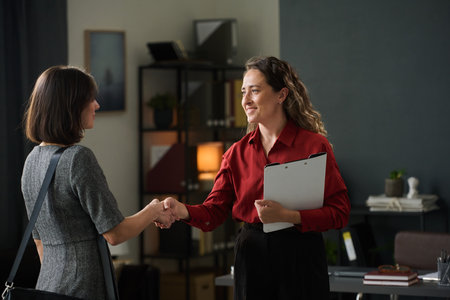 Two businesswomen greeting each other with handshake in modern office setting symbolizing professional agreement. One holding clipboard while they stand near desk and shelvesの写真素材