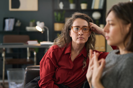 A counselor, wearing glasses and red shirt, is attentively listening to client in well-organized office. Background includes desk lamp, bookshelves, and various office suppliesの写真素材